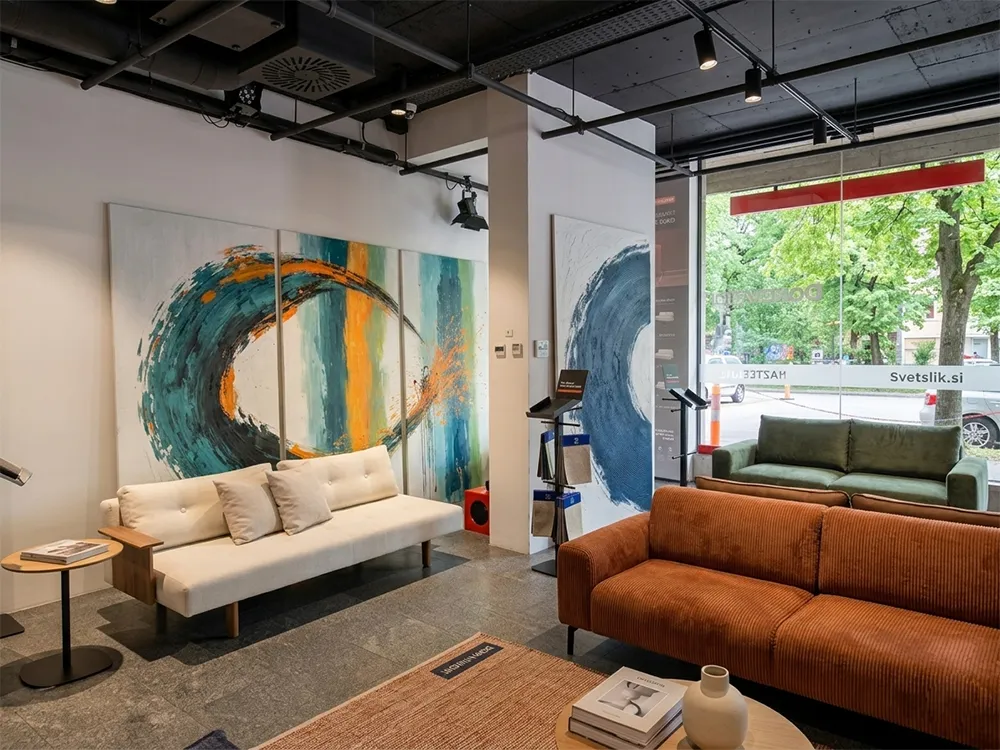 Tridelna umetniška slika triptih v teal in oranžnih tonih ob modernem raztegljivem kavču z lesenimi nasloni blagovne znamke Innovation Living v salonu Dom Ambient.