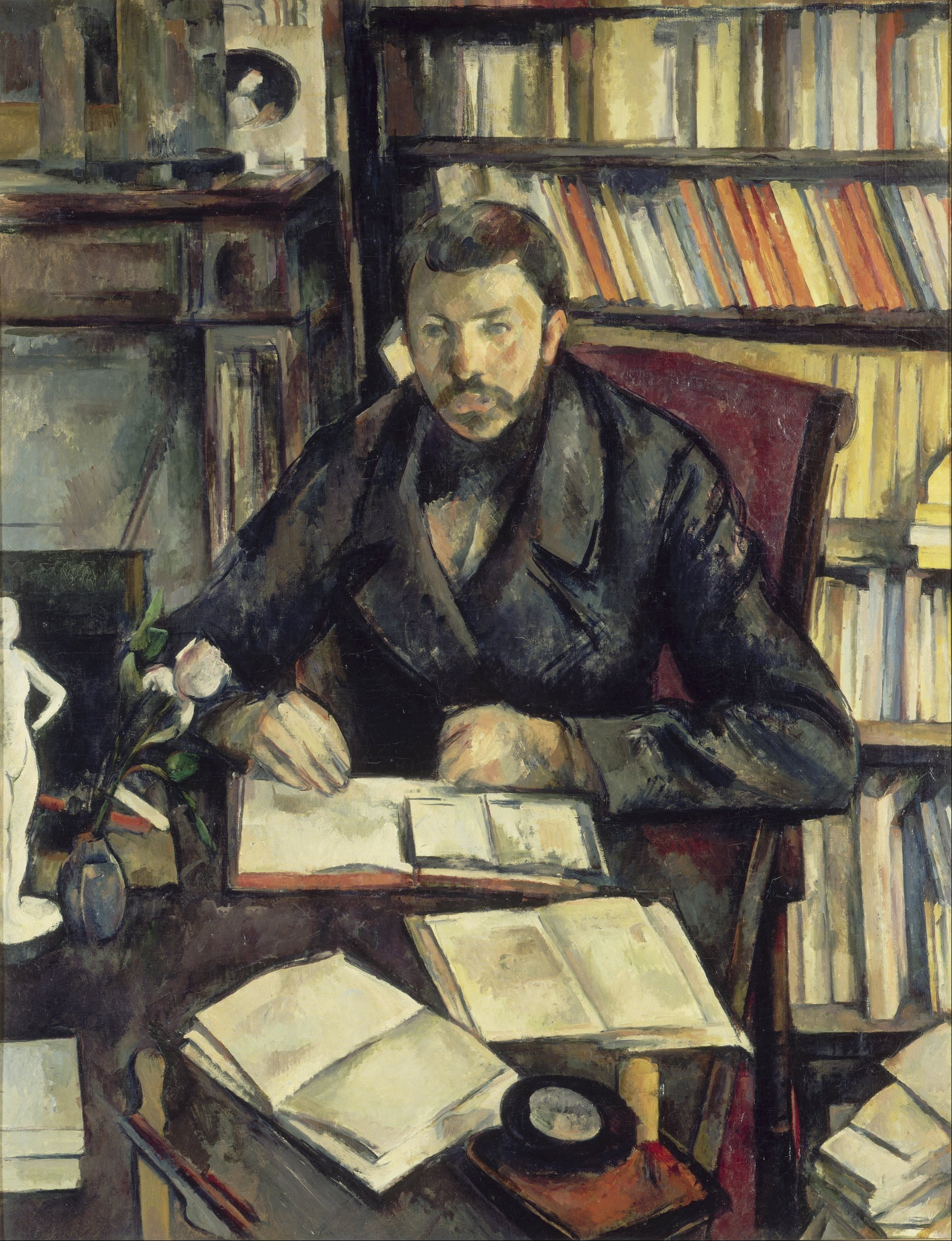 A moustached man sits at a table before a wall of bookshelves, his hands together over an open book, at the left a white plaster statuette and a white flower in a blue vase, Paul Cézanne, 1895–1896